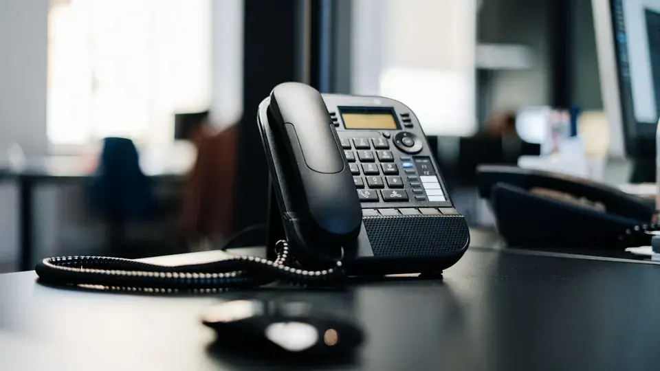 A black office desk phone with a coiled cord on a dark desk