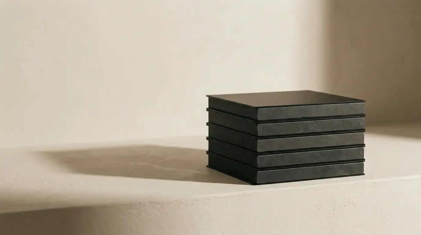 Five black-edged stone tiles stacked square on a plaster ledge in soft light