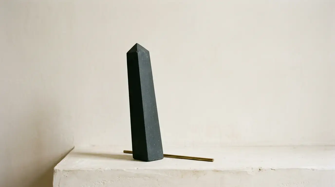 A dark tapering obelisk standing on a chalk-white plaster ledge, a brass rod lying behind it.
